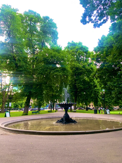 A brown iron scalloped shape fountain with a large catch basin has water spouting up. Under string lights (which are off because it’s daylight) in a city park. The skies are hazy gray and washed out. The park is lined with lush, mature tree cover.