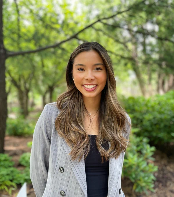 Image description: Smiling person wearing a gray pinstripe blazer, standing in a park with trees and greenery in the background.