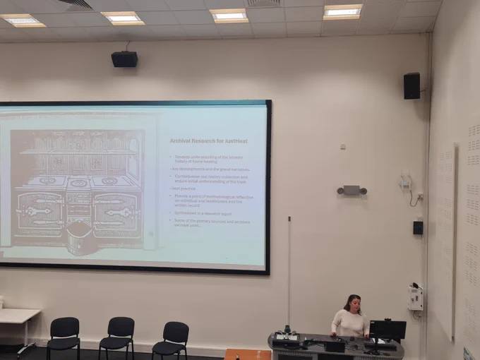 Kathy stands at the front of a lecture theatre, delivering a presentation on research methods for the closing session. She stands in front of a computer desk. In the middle of the image is a large screen. On this is information about archival research for the JUSTHEAT project.