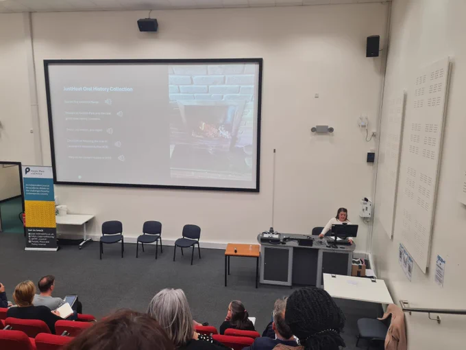 Kathy stands at the front of a lecture theatre, delivering a presentation on research methods for the closing session. She stands in front of a computer desk. In the middle of the image is a large screen. On this is a picture of a home fire and links to oral histories created as part of the JUSTHEAT project.