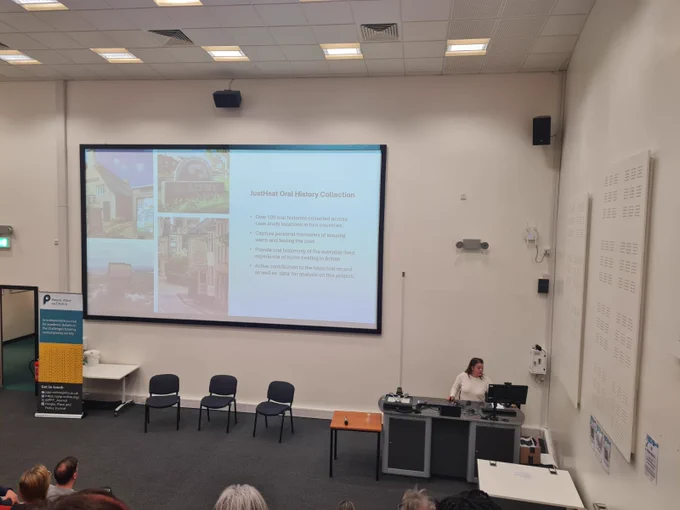 Kathy stands at the front of a lecture theatre, delivering a presentation on research methods for the closing session. She stands in front of a computer desk. In the middle of the image is a large screen. On this is information about JUSTHEAT oral history collection.