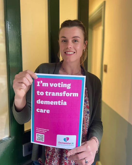 A person stands outside their front door, smiling and holding a flyer that reads, "I'm voting to transform dementia care," alongside a QR code and the logo of Dementia UK.