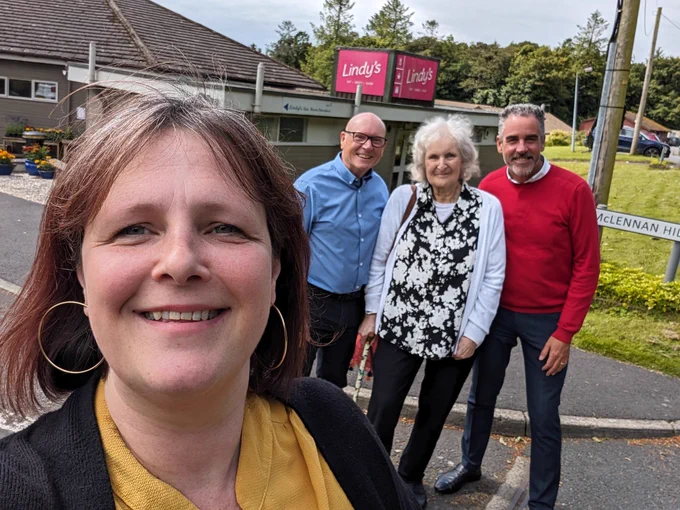 L-R me in the foreground (yellow blouse and black cardi), Gerry Lindsay CEO in a blue shirt, Blanche Nicolson MBE in a black and white floral shirt, Roddy Wright, Social Care Director in a red jumper. In the background Lindy's Tearoom, a grey building with a pink sign which was named for my Aunt, Lindy Murdoch, who was the inspiration for the charity in the 1963. The sun is shining and there is a road  sign for McLennan Hill, named after William McLennan who was an important member of the Hansel community. 