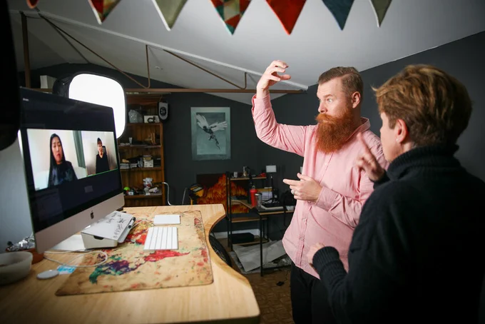 Two people are on the screen and two people are standing in an office using sign language to communicate. Pictured are Australian volunteers Ramas McRae (left) and Rebecca Ladd supporting the Mongolian Association of Sign Language Interpreters (MASLI) as Training Development Officers.