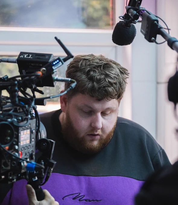 The image shows a man with curly brown hair and a full beard, wearing a purple and black sweatshirt. He appears to be concentrating, looking downward. In front of him is a professional camera setup, indicating that he is involved in a video or film production. A large microphone on a boom arm is positioned above his head. The background is blurred, but there is a window letting in some natural light. The overall scene suggests a behind-the-scenes look at a filming process.