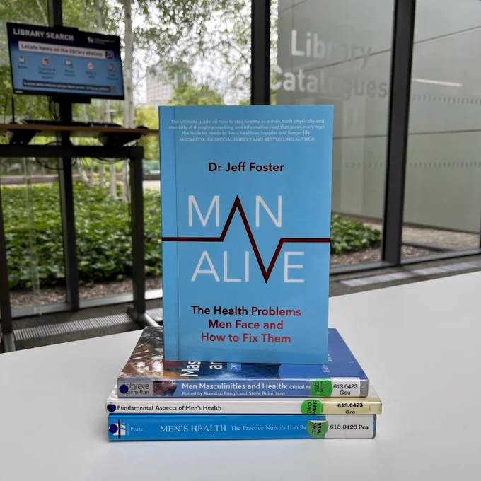 A pile of books on the subject of men's health sit on a white table with greenery in the background.