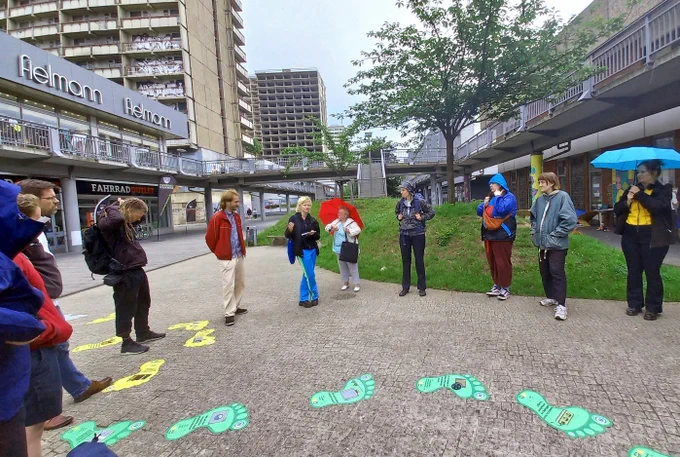 Menschen stehen im Halbkreis, im Hintergrund hohe Gebäude und eine Ladenzeile. Im Vordergrund auf dem Boden Fußabdrücke in Gelb und Grün.
