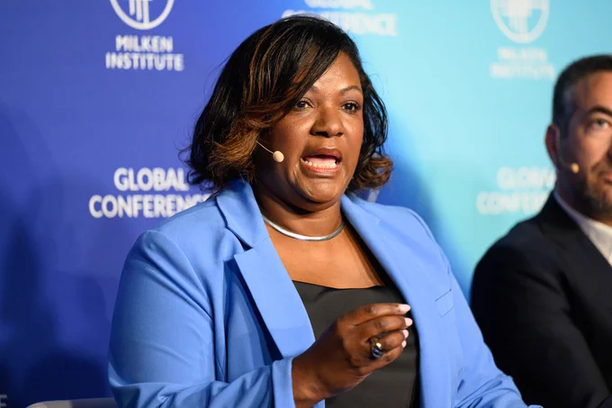 Black woman in blue blazer speaks into beige headset microphone in front of blue wall.