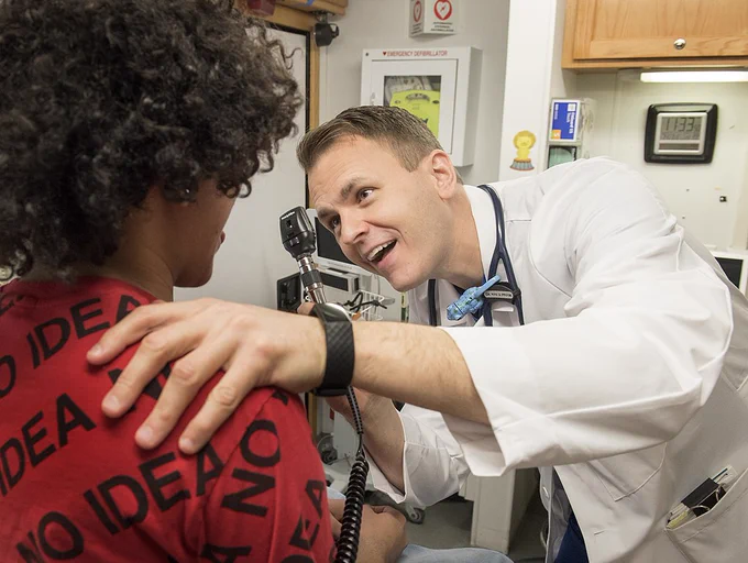 A doctor examines a patient with one hand on their shoulder and the other hand holding an instrument. 
