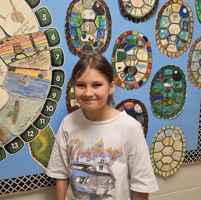 A student stands in a hallway in front of a robin blue bulletin board featuring a large turtle artwork on the left and various coloured turtle shells to the right.