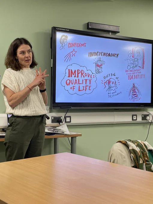 Katherine Broomfield stands to the side of a large screen showing a PowerPoint saying improves quality of life, control of my-own destiny, independence and confidence.