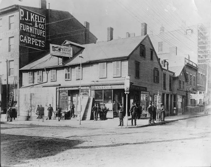  A black and white photo of a corner of two streets featuring two large buildings. One building has a painted sign on the facade reading “P.J. Kelly & Co. Furniture and Carpets.” The other building, which more resembles a house, has multiple signs. They read “Taylor Bros.”, “Bunnell’s Grand Opera House,” and the other “World’s Fair.” A group of people in period clothing are seen in front of the buildings and on the street corner.
