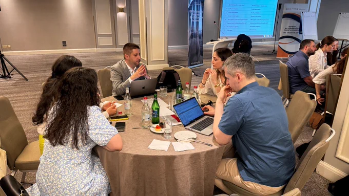 Five individuals are sat around a table in a conference room in discussion. There are laptops open, opened bottles of water and papers on the table. 