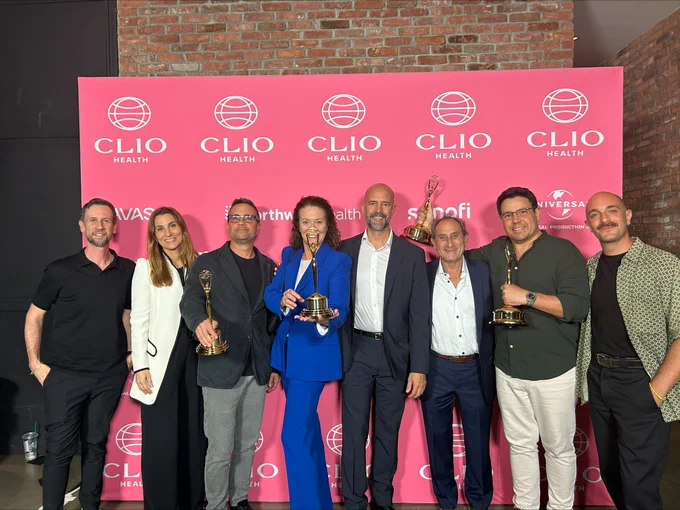 Group of eight people standing together and smiling at a Clio Health event, holding awards. They are in front of a promotional backdrop with repeated Clio Health logos. 