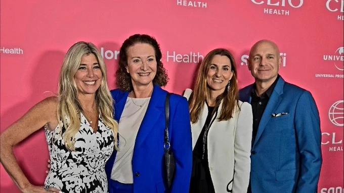 Four people standing in front of a pink Clio Health backdrop, posing for a photo. Two women on the left wear dresses, one floral and one blue, and a man on the right, wearing a blue suit. Another woman in the center is dressed in black attire.
