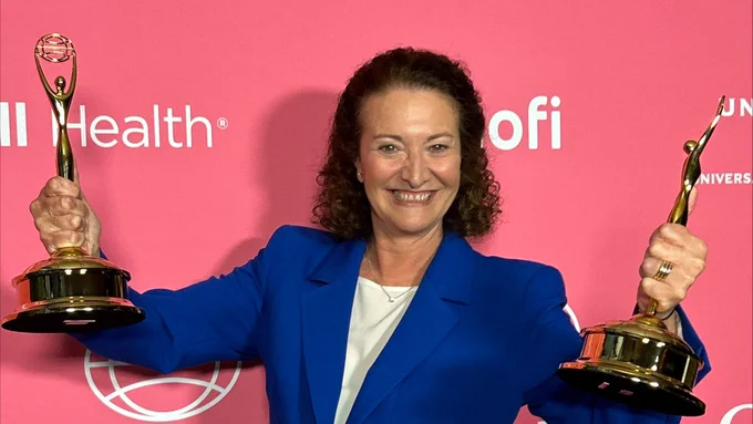 A person in a blue blazer standing in front of a pink backdrop with the text "CLIO Health" repeated. They are smiling and holding two CLIO award trophies.