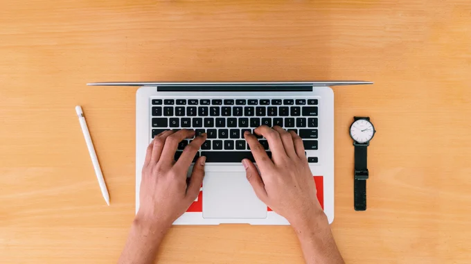 A picture of someone's hands typing on a laptop's keyboard. A pencil on the right and a watch on the left hand side of the laptop.