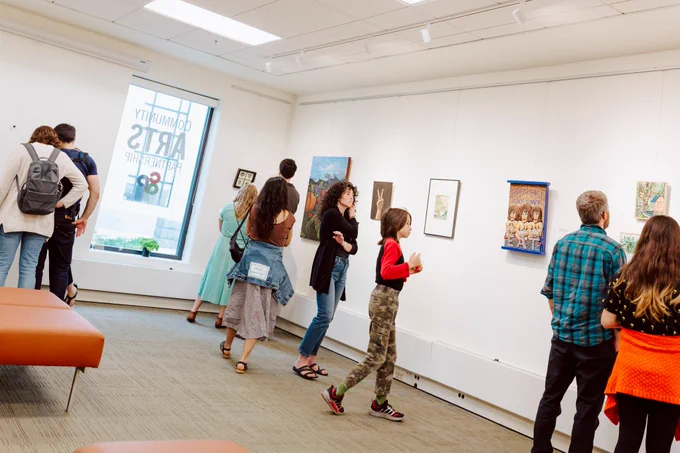 Nine people in a gallery space, with art hanging along one wall and a large window on the other.