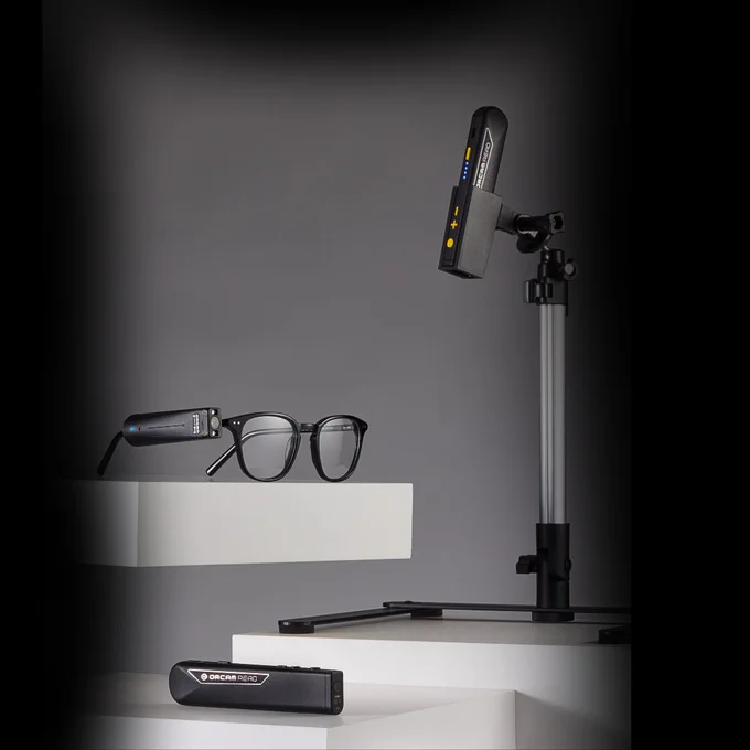 A display of Orcam's devices: the OrCam MyEye attached to a pair of black glasses, the OrCam Read, and the OrCam Read 3 mounted on a stationary stand. The devices are set against a backdrop with a black blur around them.