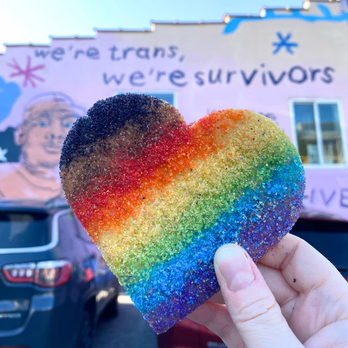 A rainbow pride heart cookie held up in front of the “We Are Universal” mural.