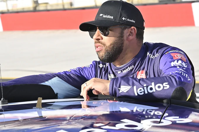 Bubba Wallace wearing the purple Leidos firesuit, a black Leidos cap and black sunglasses while leaning on the purple No 23 Camry