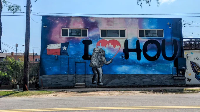 Street art in Houston. On the side of a building, art of an astronaut spray paints an "I ♥ HOU". Behind the astronaut, a Texas flag is planted into the moon's soil.