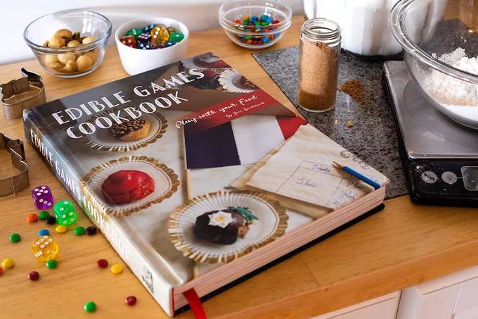 The Edible Games Cookbook closed on a table surrounded by dice, candy, and cooking supplies.