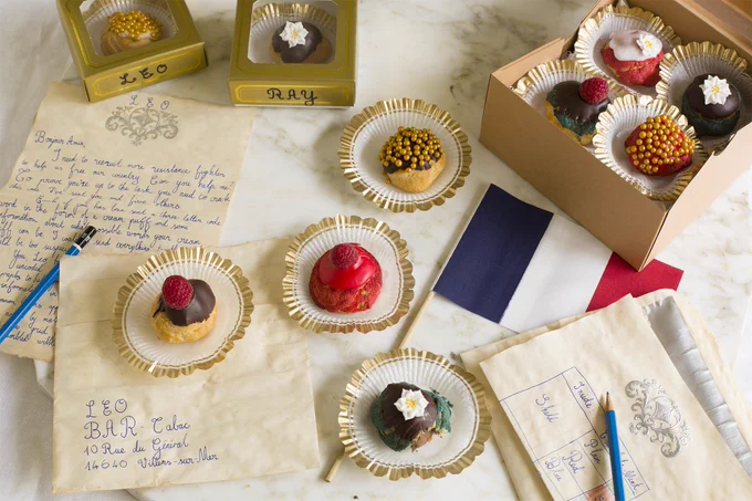 Cream puffs in boxes and on small paper holders. A French flag, pieces of paper and pencils are also on the table.