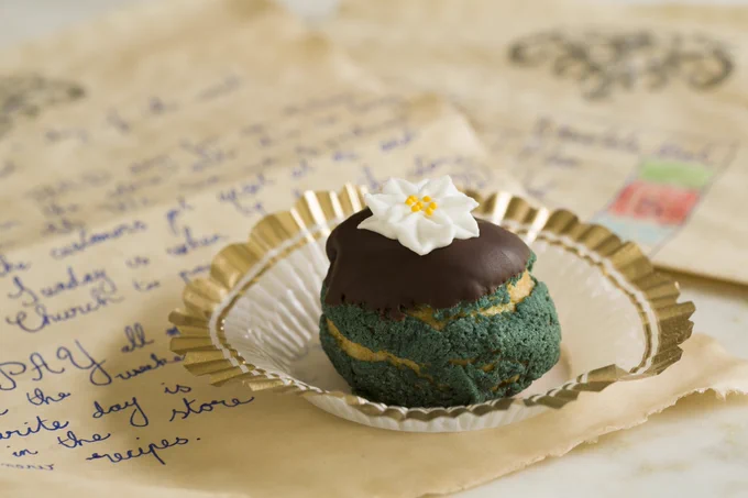 A single cream puff with a blue shell, chocolate coating, and white flower. It is sitting on top of some handwritten papers.