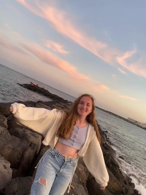 Allie wearing a beige cardigan and jeans, gesturing wide with her arms as she stands in front of the sea.