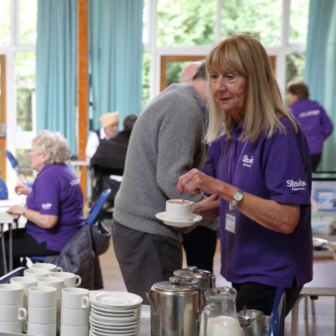 Woman collecting tea and coffee from a stall, with several people sat behind her having a chat. 