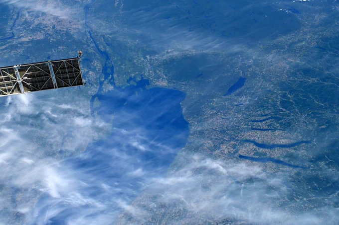 View of Syracuse and the Finger Lakes region from space, showing blue hues and wispy clouds, with part of the Space Station in the upper left corner.