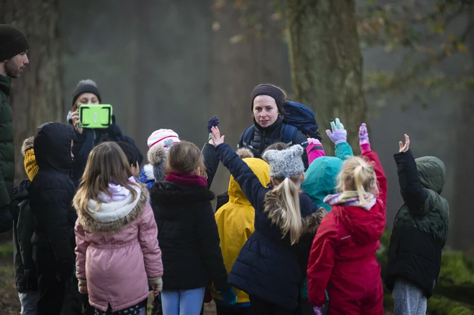 A group of children enthusiastically raising their hands in a wooded area as an adult supervises, with another adult capturing the moment on a mobile tablet.