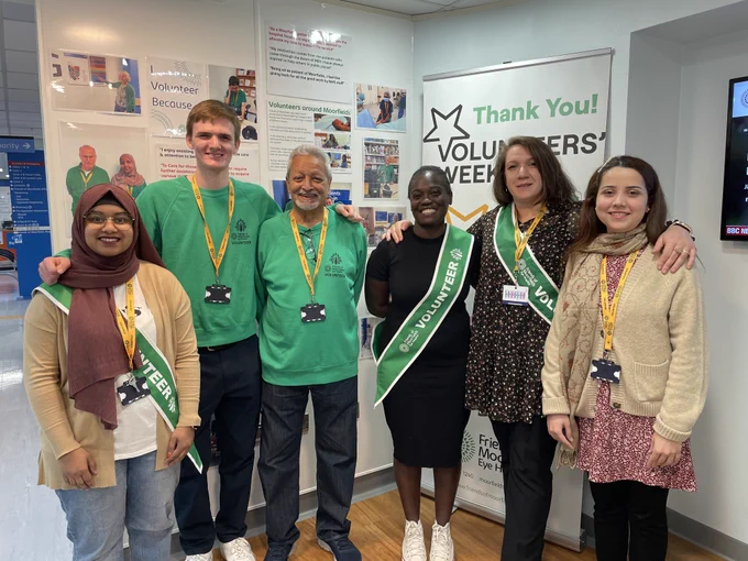 A group of six volunteers stand together, smiling, in front of a Volunteers' Week banner and display.