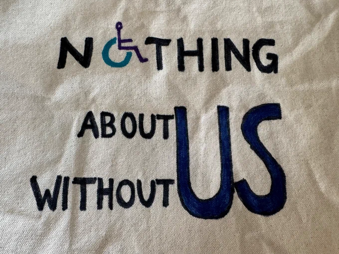 The canvas bag is hand lettered in black, blue and purple. The writing says “Nothing about US without US”. The O in “Nothing” is the wheelchair symbol in cyan with the wheelchair user drawn in purple. “US” is written once in large next to the words “about” and “without” which are placed one above the other.