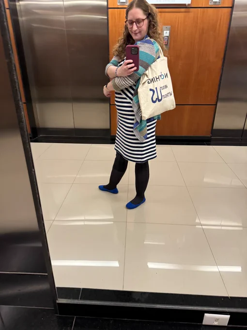 Shiri is wearing a canvas bag on which is hand lettered “nothing about us without us”. Shiri is a light skinned petite curly bronde woman wearing burgundy glasses, a blue-and-white striped dress, teal and blue cardigan, and blue flats.