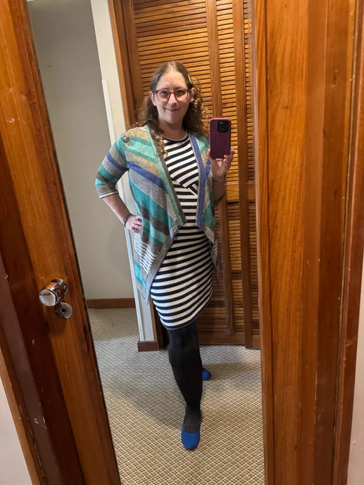 Shiri is a light skinned petite curly bronde woman wearing burgundy glasses, a blue-and-white striped dress, teal and blue cardigan, and blue flats.