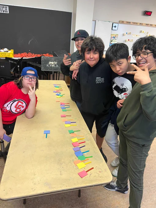 Five students stand around a table smiling towards the camera. In a line, rainbow sticks with small pieces of paper are positioned on one side of the table.