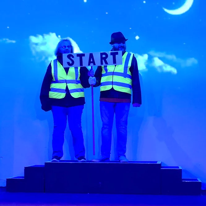 Two Progressive Players actors as race marshals in Cheshire Cats. They are holding up a large sign that says "Start".
