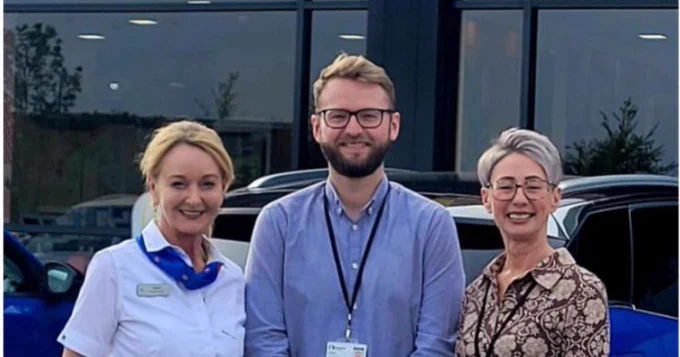 Three people standing together smiling at camera. the first person has blonde hair tied up wearing a blue shiter, neck scarf and a mcdonalds badge. the second person has short dark hear wears glasses and has a beard, they are wearing a blue shirt with a lanyard. the third peron has short blonde hair wears glasses and is wearing a partterned blouse with a lanyard.