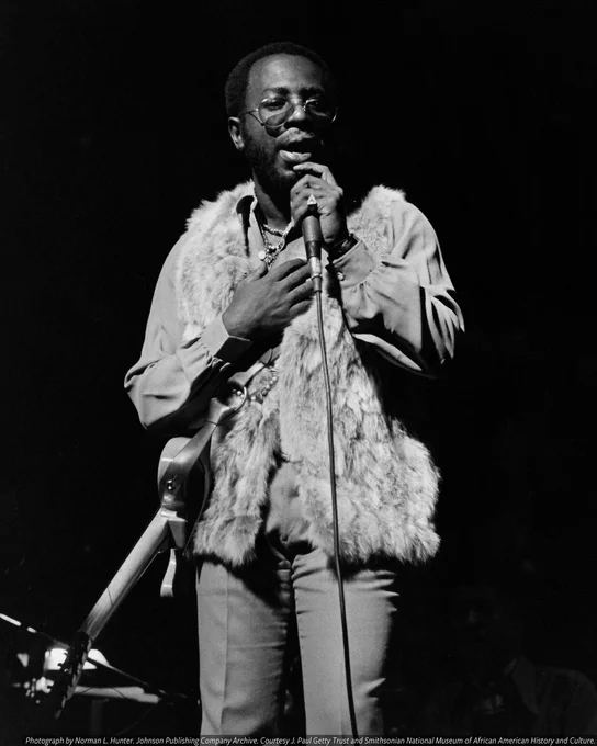 A black-and-white photo of a man (Curtis Mayfield) performing on stage. He holds a microphone to his mouth with one hand. His other hand is placed across his chest.