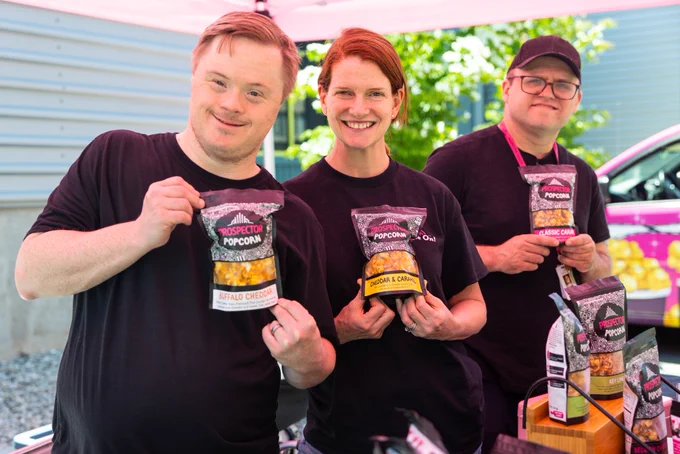 KMann, Elizabeth, and Keith smile while holding up bags of gourmet popcorn at the popup event.