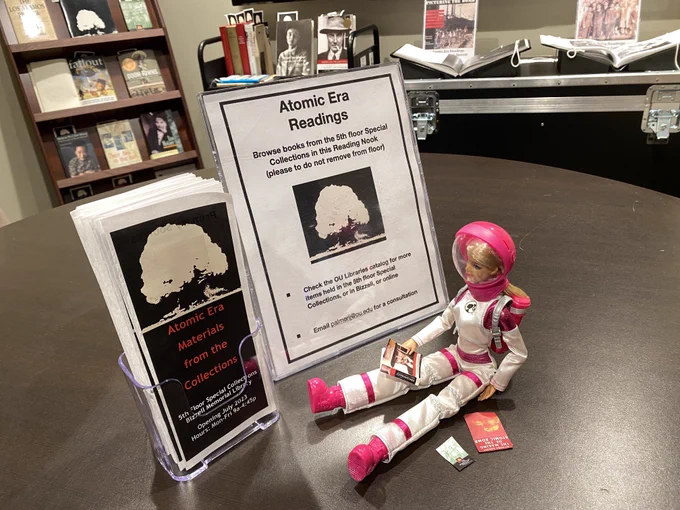Photo of astronaut Barbie doll with miniature books, near brochure and signs about the Atomic Era & Oppenheimer Exhibit.