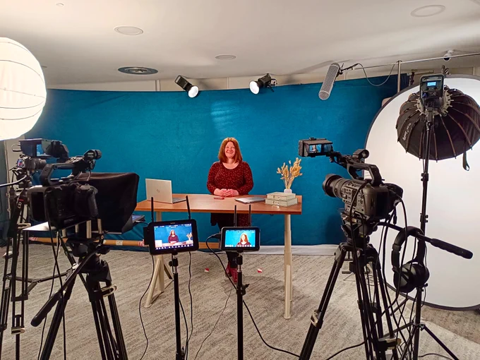Emma Darcy standing behind a desk in a studio with cameras and lights positioned around the room.