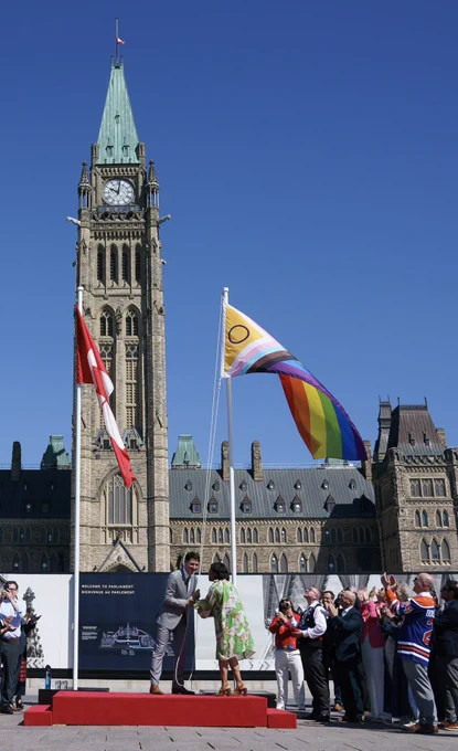 Prime Minister Justin Trudeau and Minister Marci Ien raise the Pride Flag during a ceremony on Parliament Hill. Caucus members and guests look on, applauding.