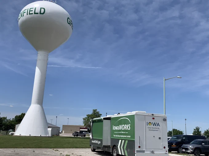 A photograph of the IowaWORSK Mobile Workforce Center on-site in Greenfield, Iowa while it is parked next to the Greenfield water tower. 