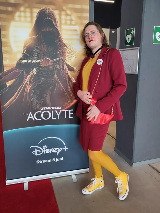 Your favourite person standing next to a standee for The Acolyte featuring Mae. I'm wearing yellow leggings, Converse, and shirt, and dark red skirt and suit jacket. Also repping a QueerSWatch pin, of course.

I'm very overdressed for what was evidently a black shirt event.