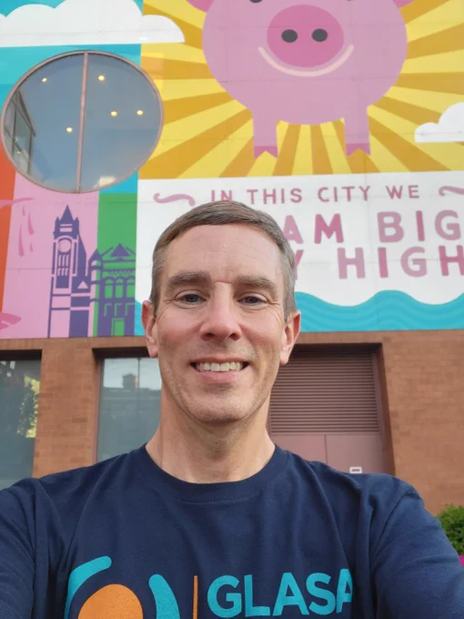 A man wearing a navy t-shirt that says, "GLASA," stands in front of a mural showing a pig with wings. Underneath the pig is text that says, "In this city we dream big & fly high."