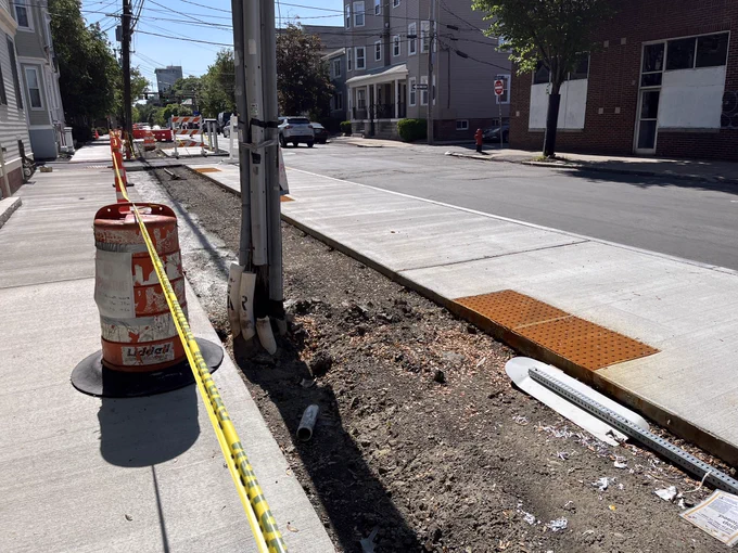 Cycle track is under construction as direct beside newly-paved sidewalk and a floating bus stop. The bus stop sign is laying on the ground. A telephone pole sites within the future bike lane.