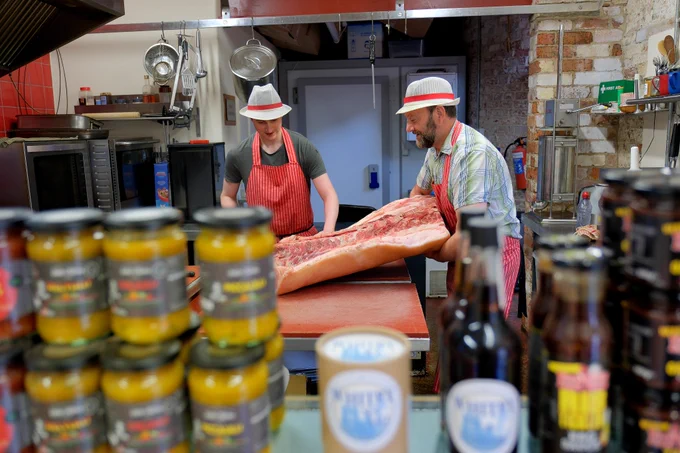 Local butchers Food2Remember in the Talbot Yard, Malton is run by a father and son team who are preparing meat in the preparation area of the butcher's shop. They also sell jars of pickles, sauces and chutneys which are pictured in the foreground of the image. 